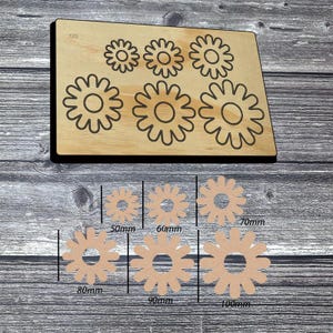 May include: A rectangular wooden cutting die featuring six flower-shaped outlines, accompanied by six corresponding tan flower-shaped cutouts. The cutouts are labeled with measurements of 50mm, 60mm, 70mm, 80mm, 90mm, and 100mm, indicating their respective diameters.