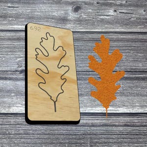 May include: A wooden die-cut tool with a black outline of an oak leaf and the number 692. An orange felt oak leaf is next to the die-cut tool. The background is a gray wooden surface.
