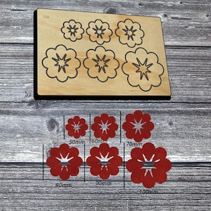 May include: A wooden die cutting board featuring six flower-shaped outlines labeled 728, paired with six red flower-shaped cutouts in sizes 50mm, 60mm, 70mm, 80mm, 90mm, and 100mm. Each flower shape features a central star-like cutout design.