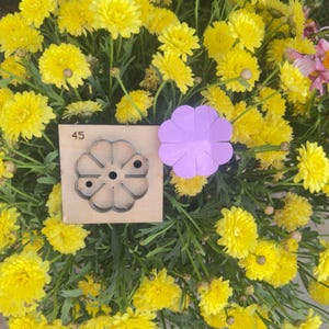 Op de afbeelding: Een houten stansgereedschap met een bloemenontwerp en het nummer 45, naast een paarse papieren bloem. De objecten staan tegen een achtergrond van levendige gele chrysanten en groen gebladerte. Het stansgereedschap wordt waarschijnlijk gebruikt voor knutselen.