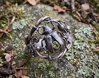 May include: A silver ring featuring a detailed beetle at its centre, accented with a small, round, light green gemstone. The band is crafted to resemble twisted vines and foliage, creating a circular frame around the central insect figure.