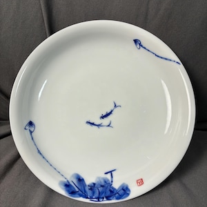 May include: White ceramic plate with a blue and white painted design. The plate features a stylized aquatic scene with two fish and aquatic plants. A small red stamp is in the lower right corner. The plate has a simple, elegant design.