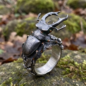 Può includere: Un anello in argento con una scultura dettagliata di un cervo volante sulla parte superiore. La fascia è lavorata per somigliare a un ramo d'albero ed è decorata con piccole foglie di quercia e ghiande scolpite. Il metallo ha una finitura scura.