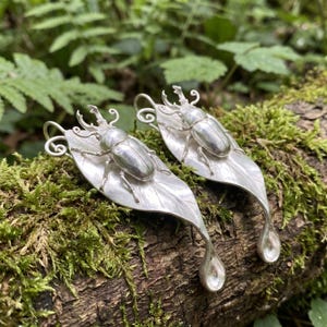 May include: A pair of silver earrings featuring a detailed stag beetle resting on a smooth leaf shape. Each earring includes a curved wire hook at the top and a small, rounded drop element at the bottom, crafted with a polished metallic finish.
