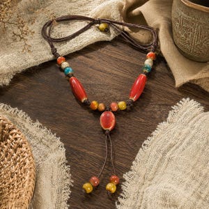 May include: A necklace featuring a dark brown cord with a series of ceramic beads in red, yellow, blue, and speckled white. The design includes two elongated red beads, a central oval red bead, and two dangling cord ends finished with small beads.