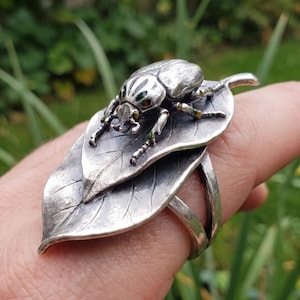 May include: A silver ring featuring a detailed beetle resting on two layered, textured silver leaves. The beetle has small green and yellow enamel accents on its head and legs, with a polished metallic finish throughout the entire piece.