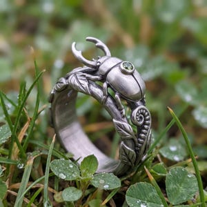 Puede incluir: Un anillo de tono plateado con un diseño detallado de escarabajo ciervo y una pequeña piedra preciosa redonda de color verde en el lomo. La banda está esculpida como una enredadera con hojas en relieve y un helecho rizado, acabado oxidado.