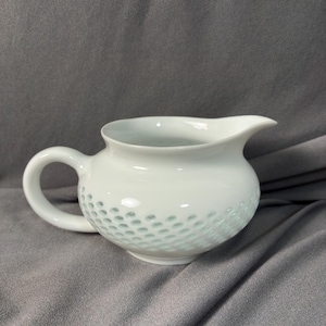 May include: A white ceramic creamer with a rounded body, a handle, and a spout. The lower section of the creamer has a textured pattern of small, evenly spaced indentations. The creamer is set against a grey background.