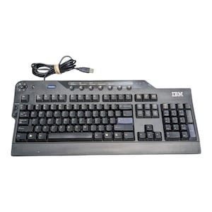 Könnte beinhalten: Eine schwarze IBM Computertastatur mit QWERTY-Layout, Ziffernblock und USB-Kabel. Die Tastatur verfügt über eine Reihe runder Mediensteuerungstasten am oberen Rand, Navigationstasten auf der linken Seite und das IBM-Logo oben rechts.