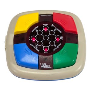 May include: A square Mini Wizard electronic game console with a beige outer casing. It features four large, colourful buttons in yellow, red, green, and blue. The centre panel displays a grid of circular lights, some illuminated red, with text including Match Me, Hot Corners, Music Maker, and Break Out.