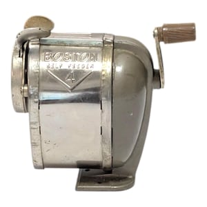 Może przedstawiać: Temperówka vintage Boston Self Feeder 4 z metaliczną srebrną płytką przednią i szarym korpusem. Urządzenie posiada ręczną korbkę z brązowym pokrętłem z boku oraz okrągły otwór po lewej stronie przeznaczony do wkładania ołówków.
