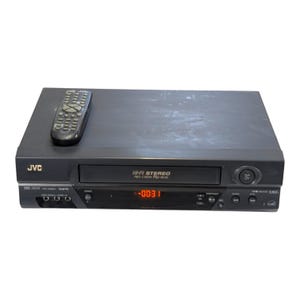 Könnte beinhalten: Ein schwarzer JVC Hi-Fi Stereo VHS-Player mit einer Digitalanzeige, die -00:31 anzeigt, einem Frontlader-Kassettenschacht und verschiedenen Bedientasten. Eine passende schwarze Fernbedienung mit zahlreichen Tasten liegt auf dem Gerät.