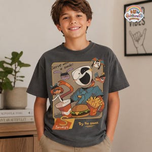 Op de afbeelding: Een antracietgrijs T-shirt met korte mouwen en een grafische print van een gemaskerd stripfiguur, een burger, frietjes en een drankje. De tekst luidt "Serve with a smile!" en "Spudsy's Try the sauce." Het ontwerp bevat diverse kleine gezichtjes.