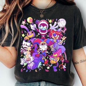 May include: Black t-shirt featuring a colorful cartoon graphic of several characters with exaggerated features. The design includes figures in purple, red, and yellow, with stars and confetti elements.