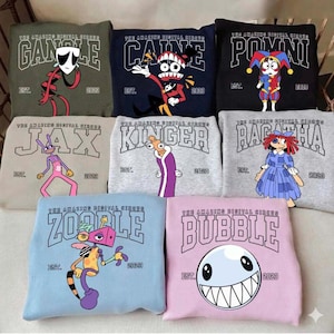 May include: A collection of sweatshirts featuring cartoon characters and text from "The Amazing Digital Circus." Each sweatshirt displays a character's name in bold letters, the year 2023, and the word "EST." Colors include olive green, navy, black, tan, grey, and light blue.