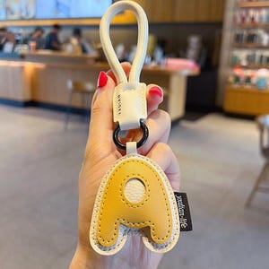 May include: Yellow leather keychain with the letter "A" and a white border. The keychain has a white loop and a black ring. The tag reads "yadian-life".