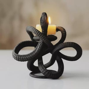 May include: A matte black candle holder shaped like a coiled snake with textured scales. The snake's head rises vertically in the centre, supporting a small, clear glass cup containing a lit cream-coloured tea light candle with a warm yellow flame.