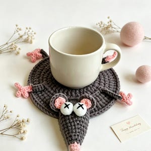 Puede incluir: Un posavasos de ganchillo gris oscuro con forma de ratón y detalles rosas en orejas, nariz y pies. El ratón tiene dos ojos blancos con pupilas negras en forma de X. Una taza de cerámica de color crema está colocada sobre el posavasos.