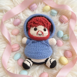 May include: A handmade crochet doll with a blue hooded sweatshirt, red hair, and a red beard. The doll is surrounded by pastel macarons and a pink ribbon on a fluffy white surface. The doll has black and white striped shoes.