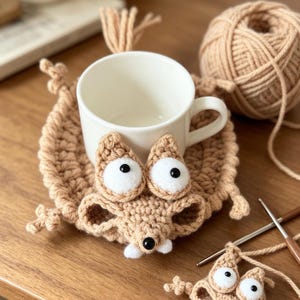 May include: A light brown crocheted coaster shaped like a mouse with large white eyes, black pupils, and two small white teeth. A white ceramic mug sits on the coaster, which features a small tail and paws. A ball of yarn and a crochet hook are nearby.