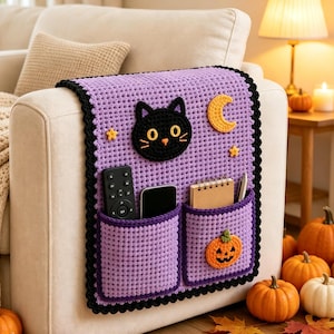 Könnte beinhalten: Ein lavendelfarbener Häkel-Armlehnen-Organizer für Sofas mit einem schwarzen Katzengesicht, einem gelben Halbmond und zwei kleinen Sternen. Er hat zwei Taschen mit Fernbedienung, Smartphone, Notizbuch und Stift. Ein kleiner orangefarbener Kürbis ist befestigt.