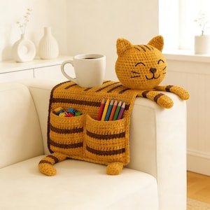 Può includere: Organizzatore per bracciolo del divano a forma di gatto, realizzato all'uncinetto, nei toni del giallo e del marrone. Presenta due tasche, una faccia di gatto e una coda. Una tazza bianca è appoggiata sopra e le tasche contengono matite e giocattoli.