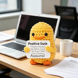 May include: A yellow crocheted duck figurine holding a white sign that reads: Positive Duck, I may be a tiny duck, but I believe in you! Go do your thing! You're Ducking Awesome! The duck has black eyes and orange feet and a small orange beak.