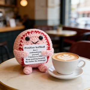 K&ouml;nnte beinhalten: Ein geh&auml;kelter rosa Softball mit roten N&auml;hten und schwarzen Augen h&auml;lt ein Schild mit motivierendem Text. Eine wei&szlig;e Tasse Kaffee mit Latte Art steht auf einer Untertasse neben dem Softball. Das Schild lautet "Positive Softball."