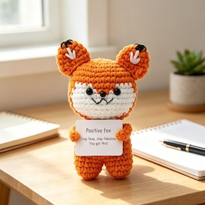 May include: A small, orange and white crocheted fox plush toy with black bead eyes and a stitched mouth. The fox holds a white rectangular sign that reads: Positive fox, Stay foxy, stay fabulous, You got this! The toy has pointed ears.