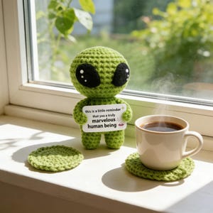 Könnte beinhalten: Eine grüne gehäkelte Alien-Puppe mit großen schwarzen Augen hält ein Schild mit der Aufschrift: „Dies ist eine kleine Erinnerung daran, dass du ein wirklich wunderbarer Mensch bist.“ Eine Tasse Kaffee steht auf einem grünen gehäkelten Untersetzer.