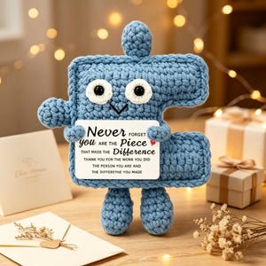 May include: A blue crocheted puzzle piece character with large white eyes and a black stitched mouth. It holds a white rectangular card with the text: Never forget you are the piece that made the difference. Thank you for the work you did.