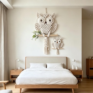 May include: A bedroom scene featuring a large macrame owl wall hanging with a smaller owl below. The owls are made of cream-colored rope and are mounted on a light-colored wall. The bed has a light-colored headboard and white bedding.