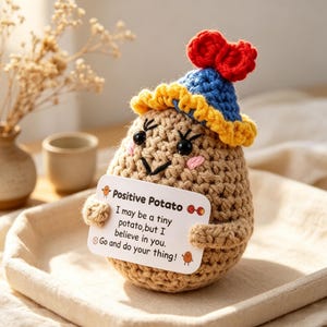 May include: A tan crocheted potato figure wearing a blue hat with a yellow brim and a red bow on top. The figure has black bead eyes, stitched eyebrows, and pink cheeks. It holds a white sign that reads: Positive Potato, I may be a tiny potato, but I believe in you. Go and do your thing!