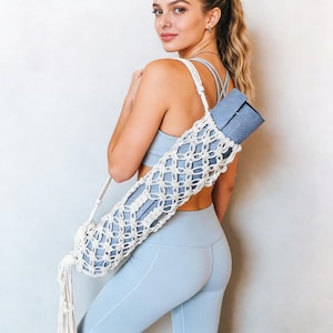 Puede incluir: Una esterilla de yoga azul enrollada se lleva en un portamantas de macramé color crema con una correa para el hombro. El portamantas tiene un diseño de red tejida y una borla en la parte inferior. Una persona lleva la esterilla de yoga y el portamantas.