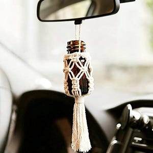 Könnte beinhalten: Eine braune Glasflasche, die am Rückspiegel eines Autos hängt, umhüllt von einer cremefarbenen Makramee-Halterung mit Quaste. Die Flasche wird von einer dünnen Schnur gehalten und bildet ein dekoratives Autozubehör.