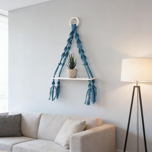 Puede incluir: Un estante de macramé para colgar en la pared con cuerda azul anudada en un patrón decorativo, suspendido de un anillo circular blanco. Una tabla de madera blanca plana sirve como estante, sosteniendo una pequeña planta verde en maceta.