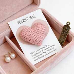 K&ouml;nnte beinhalten: Ein rosa geh&auml;keltes Herz auf einer Karte mit der Aufschrift "POCKET HUG" in einer rosa Schmuckschatulle. Die Karte hat ein herzf&ouml;rmiges Design hinter dem geh&auml;kelten Herzen. Ein Paar Perlenohrringe und ein dekoratives Metallst&uuml;ck befinden sich ebenfalls in der Box.