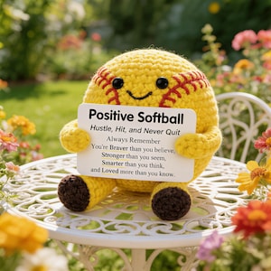 Puede incluir: Una pelota de softbol amarilla tejida con costuras rojas y una cara sonriente. Sostiene un letrero con la frase "Positive Softball" y texto inspirador. La pelota está sobre una mesa blanca, rodeada de flores.