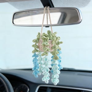 Puede incluir: Una planta colgante de ganchillo en un coche. La planta está en una maceta beige con hojas verdes y azules en cascada. La planta cuelga del espejo retrovisor con un cordón beige. Se ven el salpicadero y el volante del coche.