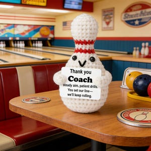 Puede incluir: Un bollo de bolos de ganchillo blanco y rojo, sosteniendo un letrero que dice "Thank you Coach". El bollo tiene ojos negros y una cara sonriente. El fondo muestra una bolera.