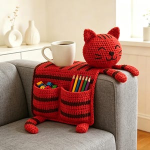 May include: A red and brown crocheted cat-shaped sofa armrest organizer. The organizer has two pockets, a cat face, and a pair of arms and legs. A white mug with coffee sits on top. The pockets contain colored pencils and small toys.