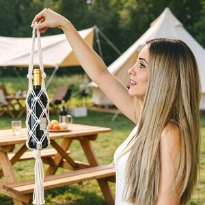 May include: A macrame wine bottle holder, crafted from off-white rope, suspends a dark wine bottle. The holder features a diamond-patterned net and a tassel. The background shows a picnic table, tent, and green grass, suggesting an outdoor setting.