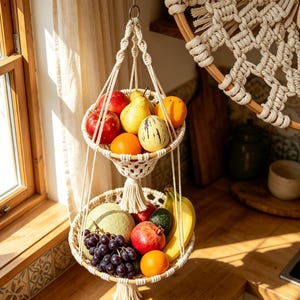 May include: A two-tiered macrame hanging fruit basket filled with fresh fruit. The top basket holds apples, pears, oranges, and a striped melon. The bottom basket contains grapes, a cantaloupe, pomegranate, avocado, banana, and an orange. The basket is suspended from a metal ring.