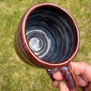 May include: A ceramic mug with a glossy, deep reddish-brown exterior and a smooth, swirling blue and grey interior glaze. The mug features a rounded, thick-walled body and a sturdy, matching handle, showcasing visible wheel-thrown texture.
