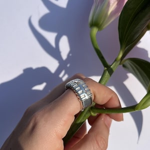 May include: A silver ring with a textured design, featuring rectangular and ridged elements. The ring is worn on a finger, held near a green stem and a pink and white flower. The background is white.