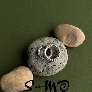 May include: Two silver rings, one with a beaded design, rest on a speckled gray stone, accompanied by two smooth, tan stones. The background is a solid, dark green. The word "S-mo" is written in black cursive.