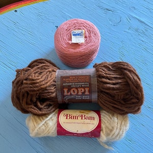May include: Three skeins of yarn, one pink, one brown, and one beige. The pink yarn is labeled "Berga" and the brown yarn is labeled "LOPI 100% pure new wool". The beige yarn is labeled "Bim Bam An unusual Irish import bulky tweed yarn".