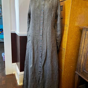 May include: A long-sleeved, floor-length dress in a dark gray textured fabric with a subtle crosshatch pattern. The garment features a high collar, a full-length front button placket with dark buttons, and vertical seam detailing along the bodice.