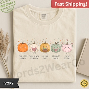 May include: An ivory t-shirt featuring a graphic of five cartoon pumpkins in orange, white, yellow, green, and pink. Each pumpkin has a face and arms, with text below reading: TAKE A DEEP BREATH, CHECK IN WITH YOUR BODY, USE YOUR TOOLS, BE KIND TO YOURSELF, YOU'VE GOT THIS.