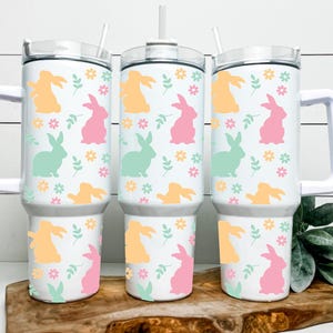 May include: Three white insulated tumblers with handles, each adorned with a repeating pattern of pastel-colored bunnies, flowers, and leaves. The tumblers have white lids and straws, perfect for cold beverages. The tumblers are approximately 10 inches tall.
